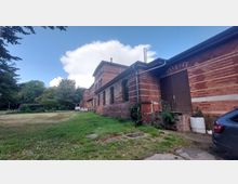 Backsteingebäude mit großen, bogenförmigen Fenstern und einer braunen Tür, umgeben von einer Grünfläche und Bäumen; im Vordergrund ein gepflasterter Weg und ein geparktes Auto. Der Himmel ist teils bewölkt.