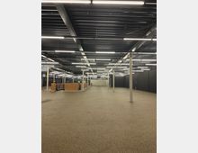 Großer, offener Innenraum mit industriellem Design, gekennzeichnet durch eine dunkle Decke mit sichtbaren Kabeln und Metallträgern sowie zahlreichen Leuchtstofflampen. Der Raum hat eine grobkörnige Bodenoberfläche, vereinzelte Säulen, schwarze Wände und eine teilweise verbaute Struktur aus Holzplatten.