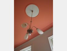 Deckenleuchte mit drei Glaslampenschirmen und einem zentralen Baldachin, befestigt an einer korallenroten Decke; im unteren Bildbereich sind Fensterrahmen erkennbar.