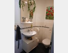 Kleines Badezimmer mit weiß gefliesten Wänden und dunklem Boden. Zu sehen sind ein Waschbecken mit Unterschrank, ein Wandspiegel mit herabhängender Kunstpflanze, eine Toilette sowie ein farbenfrohes Bild mit roten Blumen an der Wand.