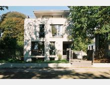 Modernes Wohnhaus mit zwei Etagen und Flachdach, umgeben von einem Metallzaun. Das Gebäude liegt in einer grünen Umgebung mit vielen Bäumen.