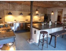 Industriell gestaltete offene Küche mit freigelegten Backsteinwänden, einer Spüle aus Edelstahl, einer zentralen Kochinsel mit dunkler Arbeitsplatte und Hocker. Die Decke besteht aus Gewölbebackstein, und hängende Metalllampen sorgen für Beleuchtung.