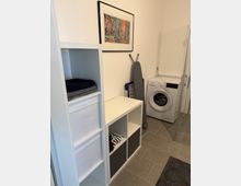 Ein Hauswirtschaftsraum mit grauen Bodenfliesen, einer weißen Waschmaschine, einem Bügelbrett, einem Regal mit Fächern für Aufbewahrung und einer abstrakten Wanddekoration. Links sind Handtücher und Stoffboxen im Regal verstaut.