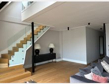 Modernes Wohninterieur mit offener Holztreppe und Glasgeländer, die zu einer Galerieebene führt. Heller Raum mit Holzfußboden, weiß-grauen Wänden, schmalen Deckenleuchten und einer schwarzen Konsole mit zwei Tischlampen.