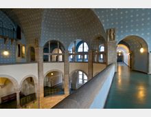 Innenansicht eines historischen Gebäudes mit gewölbten Decken, blauen Wänden mit dekorativen weißen Mustern und großen Rundbogenfenstern. Die Architektur zeigt steinerne Bögen, Säulen und Galerien mit Blick auf untere Ebenen, begleitet von einem Gang mit türkisfarbenem Boden.