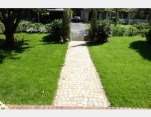 Gepflegter Garten mit grünem Rasen, durch den ein gepflasterter Weg zu einem kleinen weißen Gartentor führt. Im Hintergrund sind Einfahrten, geparkte Autos und Wohnhäuser sichtbar.