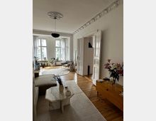 Wohnzimmer in einem Altbau mit Holzdielenboden, hohen Decken und verzierten Stuckelementen. Der Raum ist mit modernen Möbeln eingerichtet, darunter ein Couchtisch aus Marmor, ein Sideboard und diverse Zimmerpflanzen vor großen Fenstern mit dünnen Vorhängen.