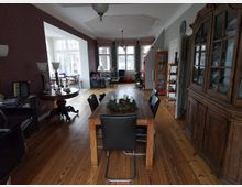 Wohnbereich eines Hauses mit Holzfußboden, der in Ess- und Sitzbereiche unterteilt ist. Im Vordergrund ein Esstisch mit schwarzen Stühlen, ein antikes Sideboard und ein runder Beistelltisch; im Hintergrund ein Sitzbereich mit großen Fenstern, die viel Tageslicht hereinlassen.