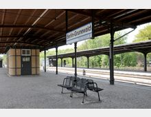 Bahnhof Berlin-Grunewald mit Bahnsteigen und Überdachung aus Holz und Stahlstützen, Sitzbänken aus Metall und einem kleinen Gebäude mit Backsteinfassade; im Hintergrund Bäume und grüne Vegetation.