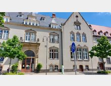 Historisches Gebäude mit heller Fassade, großen Fenstern mit steinernen Umrandungen und einem verzierten Eingang mit Rundbogen und dekorativen Elementen. Vor dem Gebäude stehen zwei Bäume sowie ein Verkehrsschild mit Fahrradsymbol.