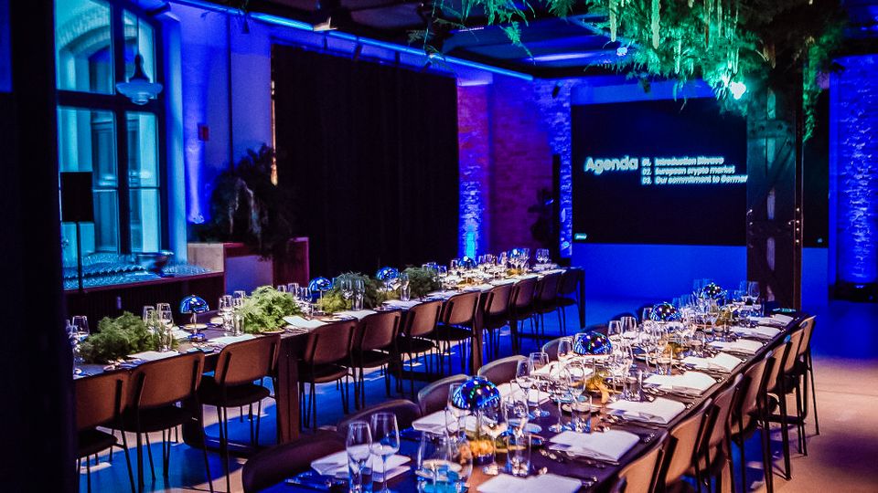 Großer Veranstaltungsraum mit langen, festlich gedeckten Tafeln und Reihen von Stühlen, in blauem Licht illuminiert. An der Decke hängen grüne Pflanzeninstallationen, im Hintergrund sind eine Projektionsfläche und hohe Fenster zu sehen.
