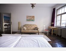 Ein schlicht eingerichtetes Zimmer mit einem Doppelbett im Vordergrund, einer gemusterten Polstercouch in der Mitte und einem kleinen Holzschreibtisch mit Stuhl rechts vor einem Fenster mit violetten Vorhängen. Im Hintergrund ist durch eine offene Tür ein angrenzender Raum mit einem Tisch und Stühlen sichtbar.