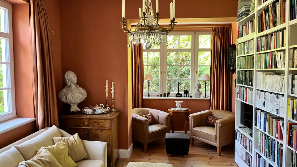 Gemütliches Wohnzimmer mit terrakottafarbenen Wänden, großem Kronleuchter und hohen Fenstern mit schweren Vorhängen. Im Raum stehen eine helle Couch, zwei Sessel mit Beistelltisch sowie eine lange, deckenhohe Bücherwand; Holzboden und ein Teppich ergänzen die Sitzecke.