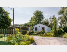 Ein kleines weißes Gebäude mit grüner Dachkante, umgeben von dichter Vegetation, steht an einer unbefestigten Wegkreuzung. Im Vordergrund befindet sich ein grüner Metallzaun mit gelben Blumen, und ein Straßenschild weist auf den „Lilienweg“ hin.