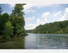 Ein ruhiger See umgeben von dichtem Wald, mit einem Uferbereich, an dem eine Person im flachen Wasser steht. Der Himmel ist teils bewölkt, und die Szenerie wirkt naturbelassen.