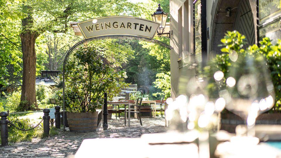 Gepflasterter Außenbereich eines Gasthauses mit Bogen-Schild „WEINGARTEN“, Tischen und Stühlen sowie großen Pflanzkübeln. Rechts eine helle Hausfassade mit Eingangsbogen und Laterne, im Hintergrund ein grüner Garten mit Bäumen.