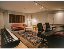 Ein modern eingerichteter Musikraum mit einem Digitalpiano im Vordergrund, einem Teppich mit detailliertem Muster, einer rosa Couch, gelbem Sessel und Lampen im Hintergrund. An der Wand befinden sich Holzpaneele und eine Tafel, umgeben von schallisolierendem Design.