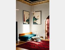 Eine stilvoll eingerichtete Ecke mit einer modernen, samtgrünen Sitzbank, einem goldfarbenen Couchtisch und zwei abstrakten Gemälden an der Wand. Der Raum weist eine hohe Decke mit einem dekorativen Stuckrand, einen roten Teppich und ein angedeutetes, gewölbtes Fenster mit roten Vorhängen auf.