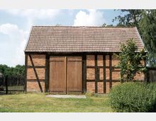 Fachwerk-Scheune mit Backsteinwänden und Holzbalken, einem Satteldach mit roten Dachziegeln und großen Holztoren in der Mitte; umgeben von Rasen, einem Zaun und vereinzelten Bäumen.