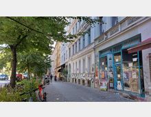 Städtische Straßenszene mit einer Reihe von historischen Wohngebäuden, deren Fassaden teils mit Graffiti und Plakaten bedeckt sind. Rechts befindet sich ein Laden mit blauen Fensterverschlägen, während auf der linken Seite ein Gehweg mit Bäumen und parkenden Fahrrädern zu sehen ist.