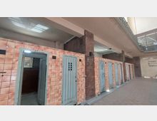 Flur mit einer Reihe von einzelnen, türartigen Kabinen mit orangefarbenen Keramikfliesen und grauen Metalltüren. Die Umgebung zeigt unverputzte Ziegelwände, eine hohe Decke mit Oberlicht und ein Geländer im oberen Bereich.