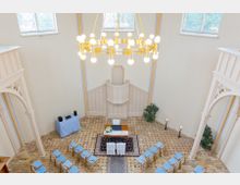 Ein heller Saal mit hohen Decken, verziert mit weißen Wänden und neugotischen Elementen. Im Mittelpunkt stehen ein geschmückter Tisch und Stühle, die auf eine Zeremonie hinweisen, umgeben von einer goldenen Kronleuchterbeleuchtung und großen Fenstern mit Bleiverglasung.