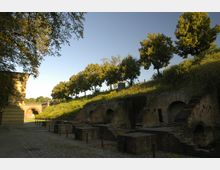 Ziegelsteinmauern einer historischen Festungsanlage mit Gewölben und Bögen, flankiert von Bäumen auf einer begrünten Anhöhe. Im Hintergrund führt ein gemauerter Torbogen über einen gepflasterten Weg.
