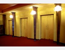 Innenbereich eines Theaters oder Kinos mit art-déco-inspiriertem Design: gelbe Wände mit floralen Mustern, Holzelemente um drei rechteckige Türen, roter Teppichboden und Wandlampen mit geschwungenen Milchglas-Schirmen.
