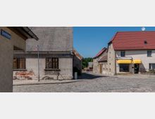 Straßenszene in einem Dorf mit Kopfsteinpflaster, umgeben von älteren Gebäuden mit Putzfassaden und roten Ziegeldächern. Links ist ein verwittertes Wohnhaus mit Holzfenstern zu sehen, rechts ein Gebäude mit gelber Markise und einem Elektro-Fachhandel.