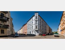 Straßenszene in einer Wohngegend mit mehrstöckigen Gebäuden im Altbaustil. Die Fassade eines Eckgebäudes ist weiß gestrichen, während die umliegenden Häuser in verschiedenen Pastellfarben gehalten sind; gepflasterte Straßen und geparkte Autos bestimmen die Umgebung.