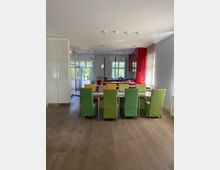 Offener Wohn- und Essbereich mit einem langen Holztisch, umgeben von Stühlen mit grünen und gelben Bezügen, im Hintergrund eine moderne Küche mit roten Schränken und großen Fenstern, die viel Tageslicht hereinlassen. Bodentiefe Türen und ein heller Holzboden verstärken den luftigen Eindruck des Raumes.