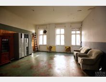 Ein einfacher Raum mit abgenutztem Boden, drei großen Fenstern und verwitterten Wänden, teils mit abblätternder Farbe. An der linken Seite stehen Metallspinde, ein Holzschrank und eine Leiter, während rechts zwei beige Sessel stehen.