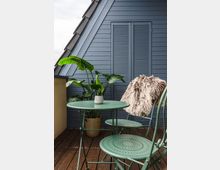 Kleine Terrasse mit Holzboden, einem runden, grünen Metalltisch und passenden Stühlen, von denen einer mit einem Fell dekoriert ist. Im Hintergrund steht eine blaue Holzwand mit geschlossenen Fensterläden und einer Zimmerpflanze in einem goldgelben Topf.