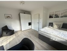 Minimalistisch eingerichtetes Schlafzimmer mit hellem Laminatboden und weißen Wänden. Zu sehen sind ein graues Polsterbett, ein großer Kleiderschrank in Weiß mit Holzoptik an den Seiten, ein schwarzer Sitzsack und an der Wand abstrakte Wandbilder sowie ein gerahmtes Foto.