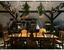 Gemütlicher Essbereich in einem Dachgeschoss mit sichtbaren schwarzen Holzbalken, großem Holztisch und verschiedenen Stühlen auf einem schwarz-weiß karierten Boden. Über dem Tisch hängen drei schwarze Pendelleuchten, Lichterketten und aufgehängtes Grün; im Hintergrund sind Fenster, Pflanzen und Dekoration zu sehen.