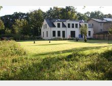 Ein weißes, modernes Wohnhaus mit schwarzen Fensterrahmen und einem dunkelgrauen Dach, umgeben von einer großen, gepflegten Rasenfläche und Blumenbeeten. Im Hintergrund sind Bäume und weitere Gebäude sichtbar.