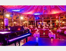 Innenraum eines festlich gestalteten Varieté-Theaters mit einem schwarzen Klavier im Vordergrund. Der Raum ist mit runden Tischen, Kerzenbeleuchtung, roten Stühlen, farbigem Baldachin und dekorativen Holzelementen ausgestattet.