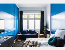 Helles Schlafzimmer mit blauen Wänden, einer großen Fensterfront mit blickdichten Vorhängen und einer dunkelblauen Couch. Im Vordergrund stehen ein Bett mit weißen Bettbezügen und dekorativen Kissen sowie ein kleiner Schreibtisch mit einer Pflanze.