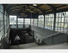 Bahnhof-Eingangsbereich mit großen Glasfenstern und Holzdecke, Treppen führen hinunter zu den Gleisen. Ein Schild weist auf die Ausgänge Schiffbauerdamm und Albrechtstraße hin, und im Hintergrund sind Fassaden von Stadtgebäuden sichtbar.