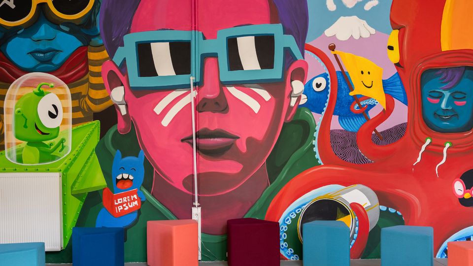 Innenraum mit einer großflächigen, bunten Wandmalerei im Pop-Art-Stil, die stilisierte Figuren und Comicmotive zeigt. Davor stehen mehrere würfelförmige Sitzhocker in verschiedenen Farben auf einem gelben Boden.