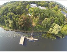 Luftaufnahme eines Sees mit einem Steg und einem angrenzenden begrünten Ufer, umgeben von dichtem Wald. Im Hintergrund sind moderne Gebäude und eine offene Rasenfläche zu sehen.