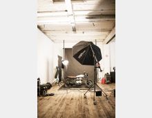 Fotostudio mit einem Motorrad als Motiv, umgeben von Beleuchtungsequipment wie Softboxen und Scheinwerfern; weiß gestrichene Wände, Holzfußboden und eine graue Hintergrundleinwand prägen den Raum.