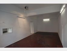 Ein leerer Raum mit weißen Wänden, einer kleinen rechteckigen Öffnung an der linken Wand und einem roten Boden mit Gebrauchsspuren. Die Decke ist schlicht, und die Beleuchtung erfolgt durch einfache Neonlampen.