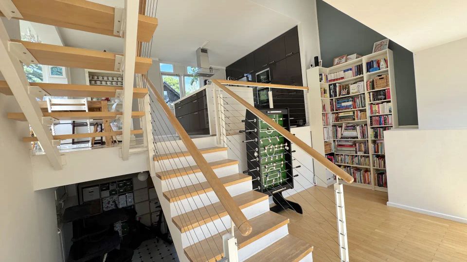 Offener Wohnbereich mit zentraler Holztreppe und Geländer aus Seilen, daneben eine moderne Küche mit dunklen Schränken. Rechts steht ein großes, weißes Bücherregal; der Raum hat helle Holzfußböden und große Fenster.