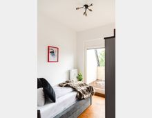Helles Schlafzimmer mit einem Einzelbett, das mit Kissen und einer Felldecke dekoriert ist, einer Balkontür, die zu einem kleinen Holzbalkon führt, und einem modernen schwarzen Deckenlicht. An der Wand hängt ein gerahmtes Bild mit rotem Rahmen, daneben steht ein kleiner, grüner Zimmerpflanzenkasten.