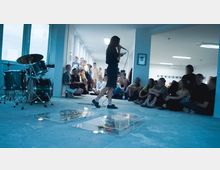 Eine Person spricht in ein Mikrofon in einem hell erleuchteten Raum mit Betonboden, umgeben von sitzenden und stehenden Zuhörern. Im Vordergrund stehen ein Schlagzeug und zwei Glasplatten mit kleinen Objekten und brennenden Kerzen darauf.