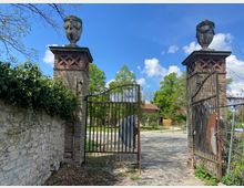 Eingangstor mit zwei gemauerten Säulen aus Ziegelsteinen, darauf steinerne Dekorationen in Form von Urnen. Ein schmiedeeisernes Doppeltor ist geöffnet, dahinter ein gepflasterter Weg, der zu Gebäuden in einer grünen Landschaft führt.