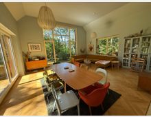 Wohnzimmer mit einem großen Holzesstisch und bunten Stühlen im Vordergrund. Der Raum verfügt über hohe Decken, große Fenster mit Blick auf einen Garten, eine lange Polsterecke, eine Bücherwand und Möbel im Retro-Stil.