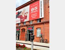 Backstein-Gebäude mit großen Fenstern und einem auffälligen Werbebanner für ein Einkaufszentrum mit 85 Geschäften. Im Vordergrund stehen eine Skulptur und rote Blumenbeete, daneben ein Fahrradständer mit einem geparkten Fahrrad.