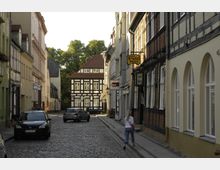 Kopfsteinpflasterstraße mit historischen Gebäuden, darunter Fachwerkhäuser und schmalen Fassaden, in einer Altstadt. Im Hintergrund steht ein auffälliges schwarz-weißes Fachwerkhaus, während links und rechts Autos parken.